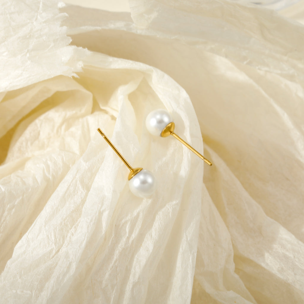 A pair of gold post earrings with white freshwater pearls, resting on a textured, crumpled cream-colored fabric background.
