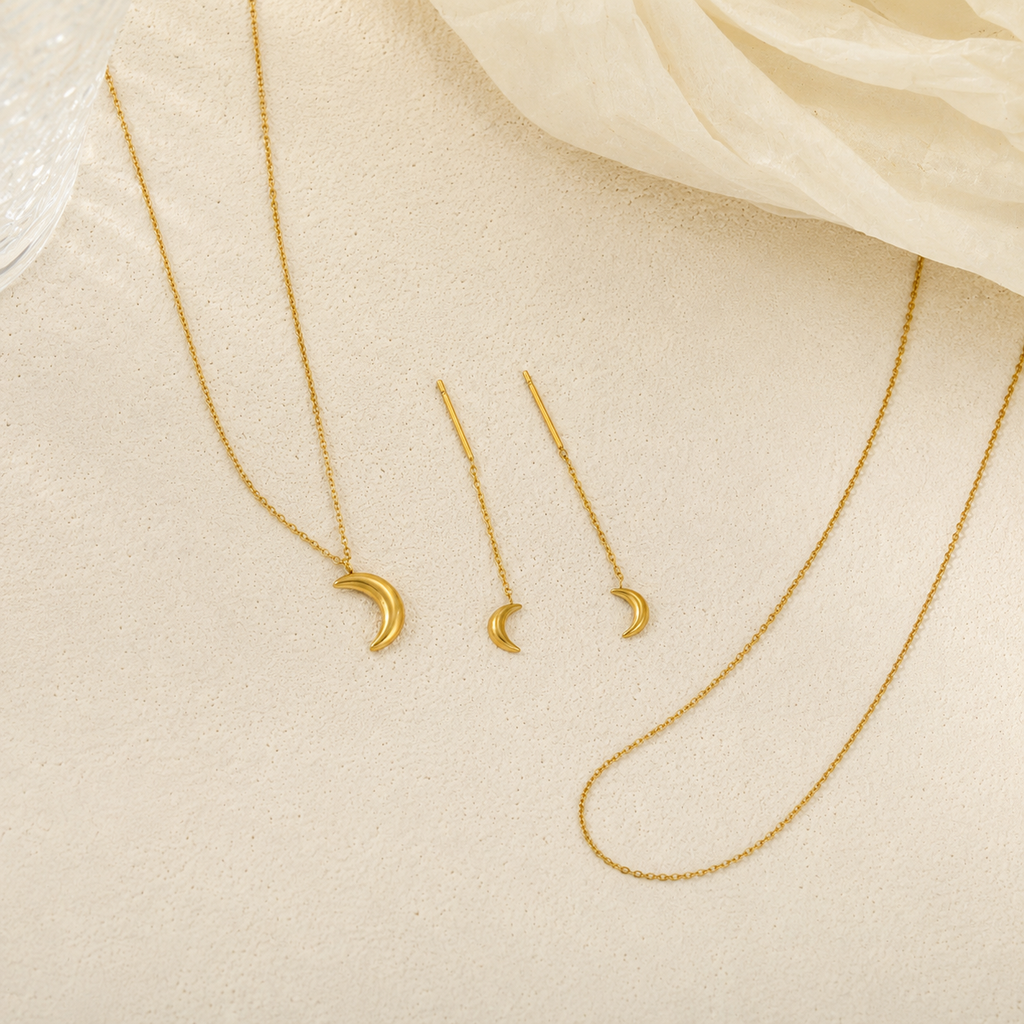 Gold jewelry set featuring a crescent moon pendant necklace, matching drop earrings, and a longer chain necklace, arranged on a textured beige surface with a soft cream fabric in the corner.
