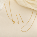Gold jewelry set featuring a crescent moon pendant necklace, matching drop earrings, and a longer chain necklace, arranged on a textured beige surface with a soft cream fabric in the corner.