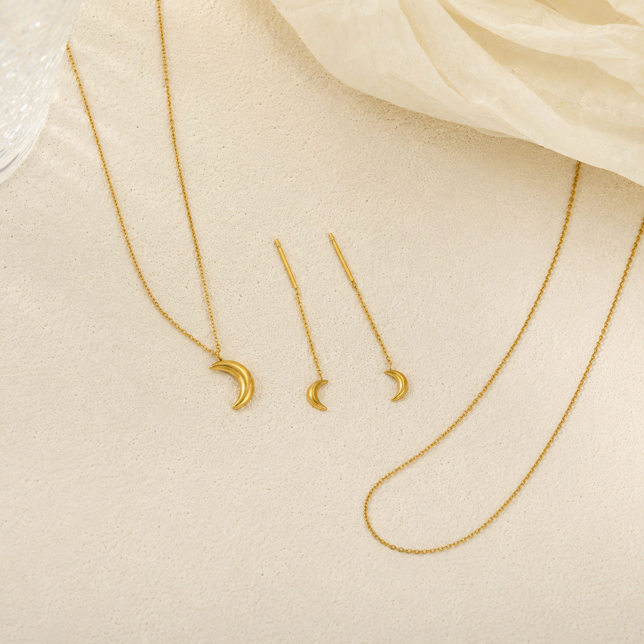 Gold jewelry set featuring a crescent moon pendant necklace, matching drop earrings, and a longer chain necklace, arranged on a textured beige surface with a soft cream fabric in the corner.